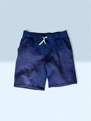 Marinblå shorts med snörning - Snygga marinblå shorts med vit snörning i midjan och två sidofickor. Shortsen har en liten röd broderad logga framtill och är tillverkade i mjuk bomull, perfekta för sommardagar.Den enda defekten med detta plagg e att de är lite solblekta i färgen