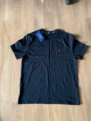 Polo Ralph lauren t-shirt svart  - Klassisk svart t-shirt från Polo Ralph Lauren med rund hals och korta ärmar. T-shirten har den ikoniska röda logotypen broderad på bröstet och är tillverkad i mjuk bomull för en skön känsla. Perfekt till jeans eller shorts.