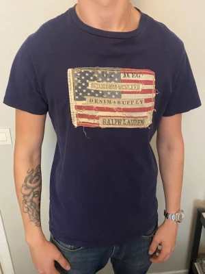 Blå Ralph Lauren t-shirt med flagga - Mörkblå t-shirt från Ralph Lauren med ett stort amerikanskt flaggtryck och texten 'Denim & Supply' på bröstet. T-shirten har korta ärmar och rund hals. Perfekt för dig som gillar en avslappnad och cool stil.