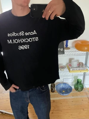 Svart longsleeve Acne Studios - Svart sweatshirt från Acne Studios med vit text 'Acne Studios STOCKHOLM 1996' på bröstet. Tröjan har avslappnad passform, rund halsringning och långa ärmar. Perfekt för en casual och trendig look.