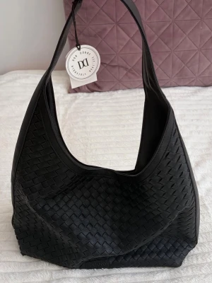 Svart flätad axelväska - Snygg svart axelväska med flätad design och mjuk struktur. Väskan har en bred axelrem och rymlig insida. Helt oanvänd, köpt på Glitter (märke ”don donna”) 