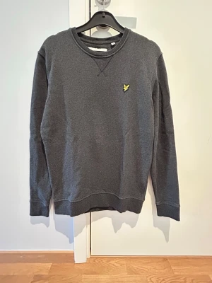Mörkgrå sweatshirt Lyle&Scott - Mörkgrå sweatshirt Lyle & Scott. Liten gul logga på bröstet. Perfekt för en avslappnad och stilren look.