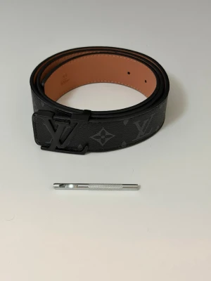 Svart bälte - Snyggt svart bälte med klassiskt monogrammönster. Bältet är gjort i skinn med brun insida och har exklusiva detaljer. Perfekt accessoar för att lyfta din outfit. 