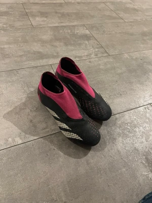 Svarta och råsa fotbollsskor - Säljer ett par stilrena fotbollsskor i svart och vinrött med vita detaljer. Skorna har en strumpliknande passform utan snörning och är tillverkade i syntetmaterial för bra grepp och kontroll på planen. Perfekta för dig som vill sticka ut på fotbollsplanen.