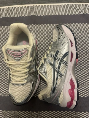 Asics skor - Snygga Asics sneakers i vitt och silver med mesh-ovandel och detaljer i rosa. Skorna har klassisk låg profil, svarta och rosa detaljer på sulan samt Asics-logga på sidan. Perfekta för dig som gillar sportig och trendig stil.