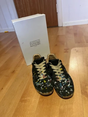 Maison Margiela svarta sneakers med färgstänk - Unika svarta sneakers från Maison Margiela med färgstänk i flera färger över hela skon. Skorna har beige snörning och detaljer samt klassisk låg siluett. Materialet är mocka och läder, vilket ger en lyxig känsla och cool look.