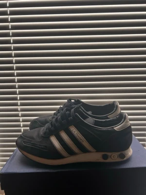 Adidas LA Trainer svarta sneakers - Klassiska Adidas LA Trainer sneakers i svart med vita och grå detaljer samt de ikoniska tre ränderna på sidan. Skorna har en låg profil och retrodesign med utbytbara pluggar i sulan. Materialet är en mix av mocka och mesh för en sportig look.