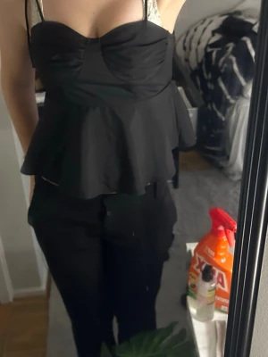 Svart peplum topp med tunna band - Snygg svart topp med tunna axelband och peplum-aktig volang nedtill. Toppen har en söt bystdetalj och sitter tight upptill. Perfekt att styla med både jeans och kjol för en trendig look.