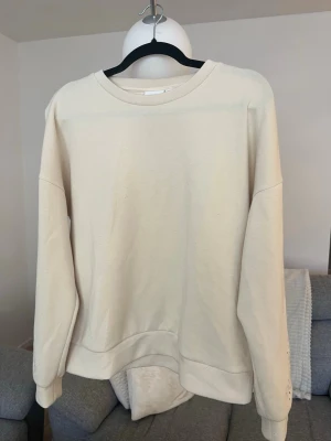 Beige sweatshirt med broderade detaljer - Säljer en beige sweatshirt i mjukt material med långa ärmar och broderade blommönster på ärmarna. Tröjan har rund halsringning och avslappnad passform. Perfekt för dig som gillar stilrena plagg med en twist.