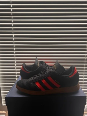 Adidas Busenitz svarta/röda sneakers - Snygga Adidas Busenitz sneakers i svart mocka med röda detaljer och klassiska tre ränder på sidan. Skorna har gummisula och svart snörning. Tungan har guldfärgad Adidas-logga och modellen har en låg profil som passar streetwear-stilen.