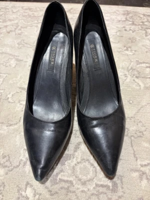Svarta pumps från Tiger of Sweden - Stilrena svarta pumps från Tiger of Sweden i äkta läder. Klassisk spetsig tå och smal klack som ger en elegant siluett. Perfekta för dig som gillar tidlös och snygg design. Passar till många olika looks.