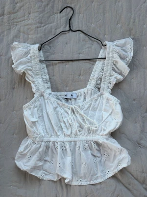 Vit broderad blus med volangärmar - Söt vit blus med broderade hålmönster och volangiga axelband. Blusen har resår i midjan och en liten knytning framtill. Perfekt för en somrig och romantisk look. Tillverkad i ett lätt och luftigt bomullstyg.