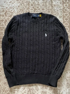 Svart kabelstickad tröja Polo Ralph Lauren - Kabelstickad svart tröja från Polo Ralph Lauren med rund halsringning och vit broderad logga på bröstet. Tröjan har ribbade muddar vid ärmslut och nederkant. Perfekt för dig som gillar klassisk och stilren design. Skulle säga att den passar i S också 