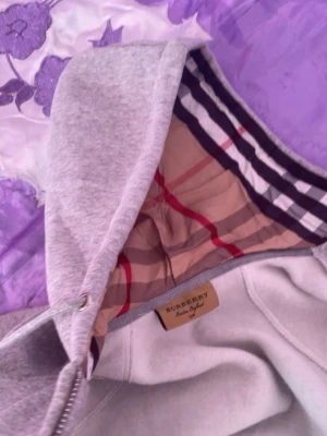 Grå hoodie från Burberry med huva - Säljer en grå hoodie från Burberry med klassiskt rutigt foder i huvan och snygga metalldetaljer på snörena. Hoodien har dragkedja och är perfekt för dig som gillar stilrena och exklusiva plagg. Passar till det mesta och har en riktigt clean look.