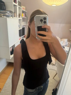 Svart linnetopp med rosetter - Snygg svart linnetopp med breda axelband och dekorativa rosetter på axlarna. Toppen har en fyrkantig ringning och är ärmlös, perfekt för en trendig och stilren look. Passar till jeans eller kjol för en cool outfit.