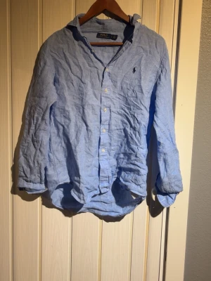 Ljusblå linneskjorta från Polo Ralph Lauren - Ljusblå skjorta i linne från Polo Ralph Lauren med klassisk krage, knäppning framtill och lång ärm. Broderad logga på bröstet. Skjortan har en avslappnad passform och är perfekt för en stilren look.