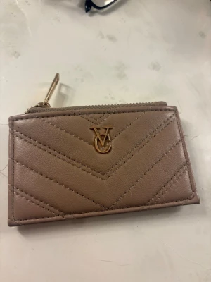  Plånbok  - Snygg beige plånbok i quiltad design med guldfärgad dragkedja och metallogo framtill. Plånboken har flera kortfack på baksidan och är tillverkad i ett material som liknar läder. Perfekt storlek för kort och kontanter.