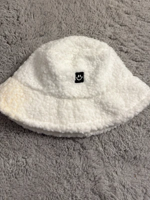Vit teddy bucket hat med smiley - Mysig vit bucket hat i teddy-material med en liten svart patch framtill som har en vit smiley. Hatten har en klassisk form med bred brätte och är perfekt för dig som vill ha en trendig och bekväm accessoar till hösten eller vintern.