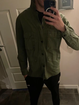 Grön overshirt med fickor - Snygg militärgrön overshirt med fyra stora fickor framtill och knäppning med svarta knappar. Overshirten har rak passform och är tillverkad i ett kraftigare bomullstyg. Perfekt att slänga över en t-shirt för en avslappnad look.