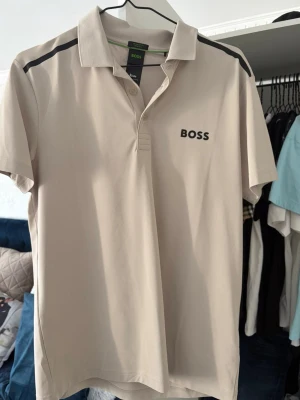 Beige kortärmad skjorta från BOSS - Snygg beige skjorta från BOSS med korta ärmar och svart logga på bröstet. Skjortan har klassisk krage, knappar framtill och svarta detaljer på axlarna. Tillverkad i stretchigt material som andas och hanterar fukt.