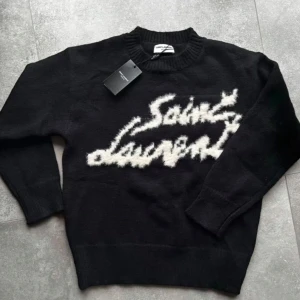 Svart stickad Saint Laurent tröja - Svart stickad tröja från Saint Laurent med vit, fluffig logotyp framtill. Tröjan har rund halsringning och långa ärmar. Perfekt för dig som vill ha en statement piece med lyxig känsla och cool design. Inga tags inga kvitton inget sånt,