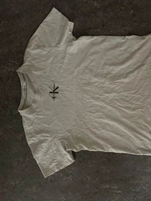 Vit basic t-shirt med tryck - En enkel vit t-shirt i bomull med korta ärmar och ett litet svart tryck på bröstet. Perfekt till jeans eller shorts för en clean och avslappnad stil.