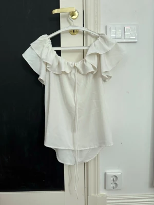 Vit offshoulder blus med volang - Söt vit offshoulder blus med bred volang runt axlarna och en lös passform. Toppen har en lätt, luftig känsla och är perfekt för varma dagar. Materialet känns mjukt och toppen har en feminin look med sin axelbandslösa design. Sitter som S och inte M/L!