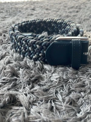 Flätat bälte från ONEWear i blått - Snyggt flätat bälte från ONEWear i olika nyanser av blått, grått och svart. Bältet har detaljer i mörkblått skinn och ett silvrigt spänne. Perfekt accessoar för att lyfta din outfit.