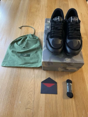 Valentino Garavani Rockrunners - Snygga sneakers från Valentino Garavani i svart och grått camomönster. Skorna har lågt skaft, svarta skosnören och detaljer i mocka och textil. Kommer med originalkartong, extra skosnören och dustbag. Perfekta för dig som gillar exklusiv streetwear.