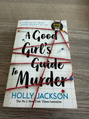 A Good Girl's Guide to Murder - En spännande pocketbok av Holly Jackson med ett omslag i vitt och svart, dekorerat med röda trådar och nålar som ger en true crime-känsla. Perfekt för dig som gillar mysterier och thrillers. Boken är på engelska och har ett modernt, ungdomligt uttryck.