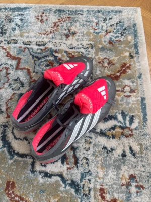 Adidas Predator fotbollsskor svart/röd - Säljer ett par Adidas Predator fotbollsskor i svart och rött med vita detaljer. Skorna har klassisk Predator-design med texten 'EST. 1994' på plösen och mönstrad sula för extra grepp. Perfekta för dig som vill sticka ut på planen.