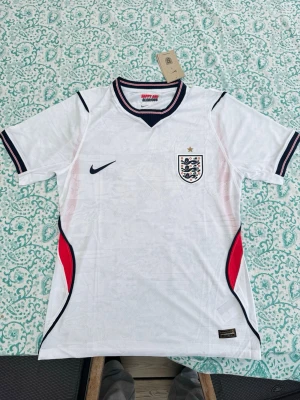 England Nike vit matchtröja herr - Säljer en vit England matchtröja från Nike med broderad landslagssköld och stjärna på bröstet. Tröjan har svarta och röda detaljer längs sidorna och på ärmsluten, samt rundad krage med röd och svart rand. Tillverkad i lätt funktionsmaterial.