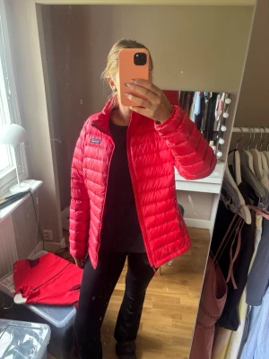 Röd dunjacka från Patagonia - Snygg röd dunjacka från Patagonia med quiltad design och hög krage. Jackan har dragkedja framtill och två sidofickor med dragkedja. Perfekt för kalla vinterdagar och riktigt lätt att bära. Klassisk logga på bröstet.