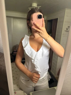 Vit ärmlös topp med volang - Säljer en snygg vit ärmlös topp med djup v-ringning och volangdetalj längs halsen. Toppen har en feminin och fräsch look, perfekt till sommaren. Passar till både jeans och kjol för en trendig outfit.