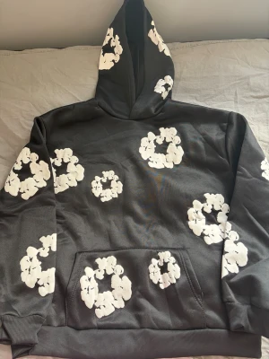 Denim Tears set - Säljer en svart hoodie och byxor från Denim Tears i storlek M med vita blommönster över hela tröjan och huvan. Hoodien har en klassisk känguruficka och ribbade muddar. Perfekt för dig som vill sticka ut med en unik och trendig look.