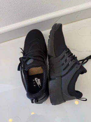 Svarta Nike Air Presto sneakers - Snygga helsvarta Nike Air Presto sneakers med låg profil. Skorna har en mesh-överdel för bra andningsförmåga, plastdetaljer på sidorna och klassisk Nike-logga på plösen och sulan. Perfekta för dig som gillar stilrena och sportiga sneakers. Använda ett par gången inga håll men lite repet sunna knappt 