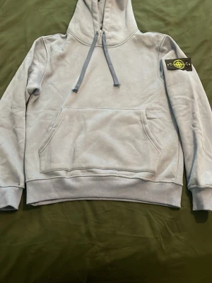 Ljusblå stone island hoodie - Snygg ljusgrå hoodie från Stone Island med klassisk patch på vänster ärm. Hoodien har huva med dragsko, stor magficka och ribbade muddar vid ärmslut och nederkant. Perfekt för en avslappnad och trendig stil.