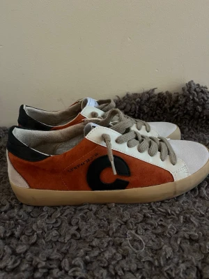 Unika suede sneakers i olika färger  - Snygga sneakers med orange mocka på sidan, vita detaljer och beige snörning. Skon har en stor svart C-logga på sidan och svart parti vid hälen. Yttersulan är beige och ovandelen är i en mix av mocka och textil. Perfekt för dig som gillar en unik och trendig look.
