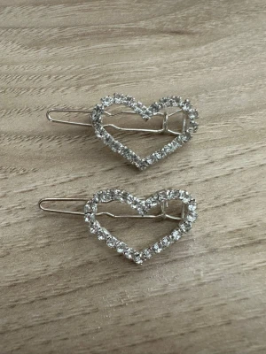 Hårspännen med hjärtan och strass - Två silvriga hårspännen i form av hjärtan, dekorerade med gnistrande strass-stenar runt hela kanten. Perfekta för att ge frisyren extra bling och en söt touch. Metallmaterial och enkel clip-on funktion.