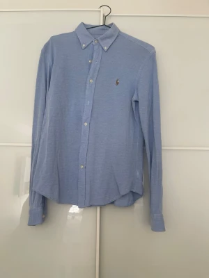 Blå slim fit skjorta från Ralph Lauren - Klassisk ljusblå skjorta från Ralph Lauren i slim fit-modell. Skjortan har knappar framtill, button down-krage och den ikoniska broderade loggan på bröstet. Tillverkad i bomull och passar perfekt till en clean och stilren look.