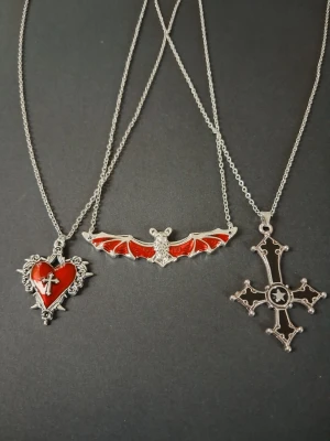 Set med tre gotiska halsband - Säljer ett set med tre halsband i silverfärgad metall. Ena har ett rött hjärta med kors och taggar, rosor det andra ett rött fladdermusmotiv och det tredje ett stort svart kors med stjärndetalj. Alla har tunna kedjor ankarlänkar och coola detaljer som passar dig som gillar alternativ stil. Materialet är av rostfritt stål 