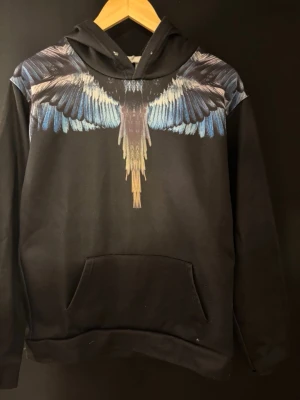 Svart hoodie med färgglada vingar marcelo burlon - Säljer en svart hoodie med stor färgglad vingdesign över axlar och bröst i blått, vitt och brunt. Hoodien har huva och en stor magficka framtill. Perfekt för dig som vill sticka ut med en unik och konstnärlig look.
