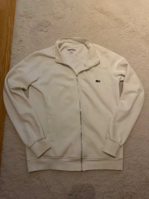 Vit ziptröja från Lacoste - Snygg vit ziptröja från Lacoste med klassisk krokodillogga på bröstet. Tröjan har hel dragkedja, ståkrage och två fickor framtill. Perfekt för en clean och sportig look. Mjuk bomullsblandning och ribbade muddar vid ärmslut och nederkant.