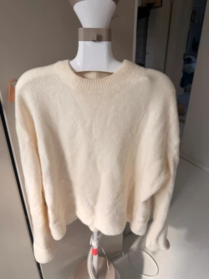 Krämvit oversized stickad tröja - Mysig krämvit stickad tröja med rund halsringning och avslappnad passform. Tröjan har långa ärmar och ribbade muddar vid ärmslut och nederkant. Perfekt för lager på lager och enkel att matcha med olika stilar.