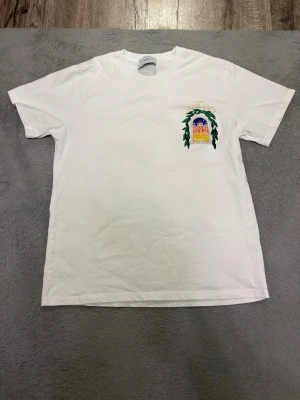 Vit t-shirt med färgglatt tryck - Vit t-shirt med färgglatt tryck på bröstet och ryggen. Trycket visar en byggnad med texten 'Avenida Casablanca' och gröna blad runt motivet. Klassisk passform och rund hals. Perfekt för dig som gillar unika prints och stilrena plagg.