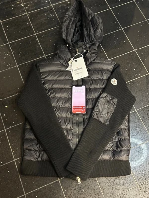 Svart pufferjacka med huva Moncler - Snygg svart pufferjacka från Moncler med stickade ärmar och vadderad kropp. Jackan har en praktisk huva, dragkedja framtill och en ficka med Moncler-logga på ärmen. Perfekt för kalla vinterdagar och riktigt trendig look.