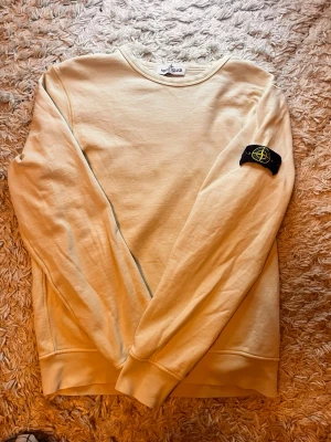 Beige sweatshirt från Stone Island - Snygg beige sweatshirt från Stone Island med klassisk rund hals och den ikoniska svarta patchen med gul logga på vänster ärm. Tröjan har en avslappnad passform och är tillverkad i mjukt bomullsmaterial. Perfekt för en clean och stilren look.