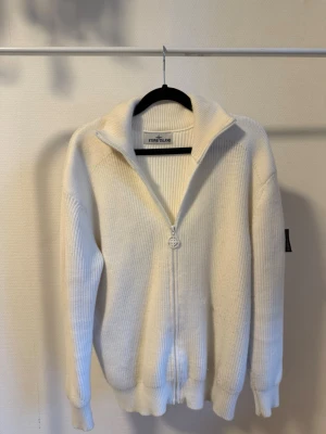 Vit stickad ziptröja Stone Island - Vit ribbstickad ziptröja från Stone Island med hög krage och dragkedja framtill. Tröjan har långa ärmar, ribbade muddar och den klassiska logotypen på ärmen. Perfekt för lager på lager och riktigt clean stil.