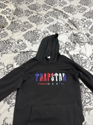 Svart hoodie med Trapstar-tryck - Svart hoodie med stor huva och trycket 'TRAPSTAR IT'S A SECRET' på bröstet i blått, rött och vitt. Klassisk känguruficka framtill och avslappnad passform. Perfekt för en streetwear-look. pris kan diskuteras 