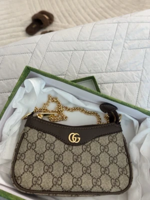 Gucci beige GG canvas handväska - Säljer en snygg beige handväska från Gucci i klassiskt GG canvasmönster med bruna läderdetaljer och guldfärgad kedja. Väskan har en elegant form och GG-logga framtill. Perfekt accessoar för dig som gillar lyxiga detaljer och trendig design.